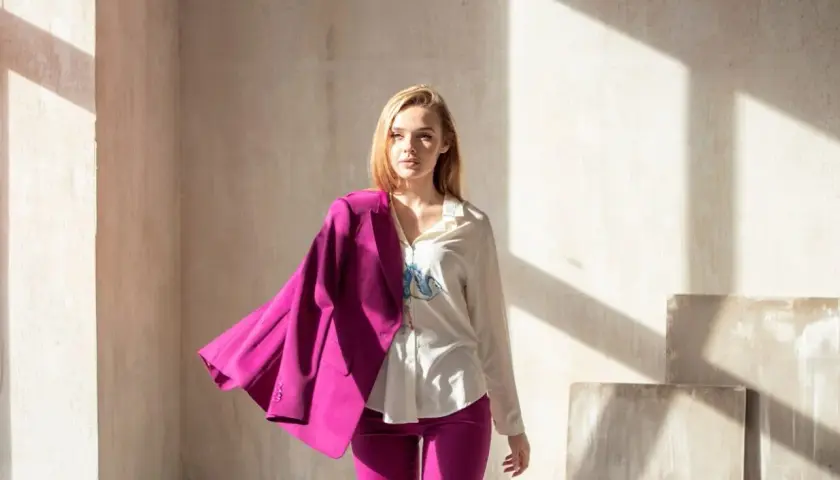 Kobieta w stylizacji viva magenta, z marynarką narzuconą na ramię, stoi w świetle słonecznym.