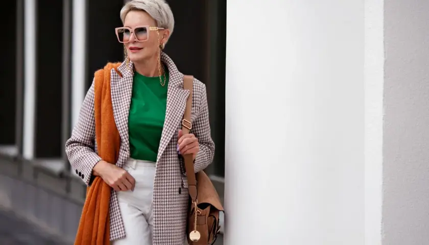 Elegancka kobieta po 60. w stylowej marynarce, zielonej bluzce i białych spodniach. Pomarańczowy szal dodaje jej uroku.