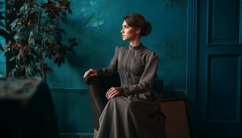 Elegancka kobieta w sukni w stylu light academia siedzi w fotelu, w tle ciemnozielona ściana i roślina.