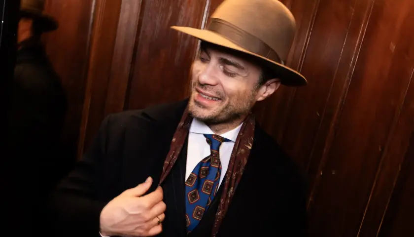 Elegancki mężczyzna w brązowym kapeluszu, czarnym płaszczu i stylowym krawacie. Pokazuje, jak nosić kapelusz męski, dodając klasy każdej stylizacji.