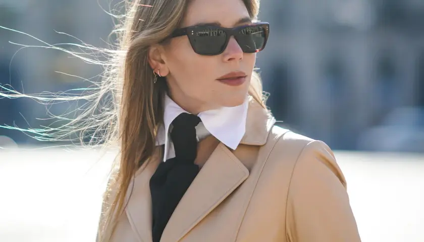 Elegancka kobieta w beżowym płaszczu, okularach przeciwsłonecznych i białej koszuli z czarną apaszką, tworzącą ciekawy rodzaj kołnierzyka.