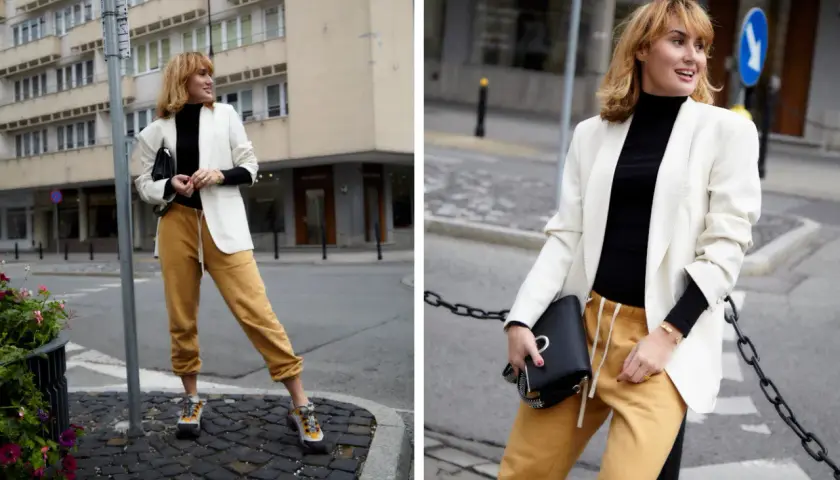 Modna kobieta w stylizacji z czarnym golfem, białej marynarce i musztardowych spodniach dresowych.