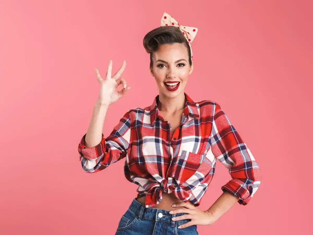 Uśmiechnięta kobieta w stylu pin-up, w kratowanej koszuli i dżinsach, pokazuje znak "OK".
