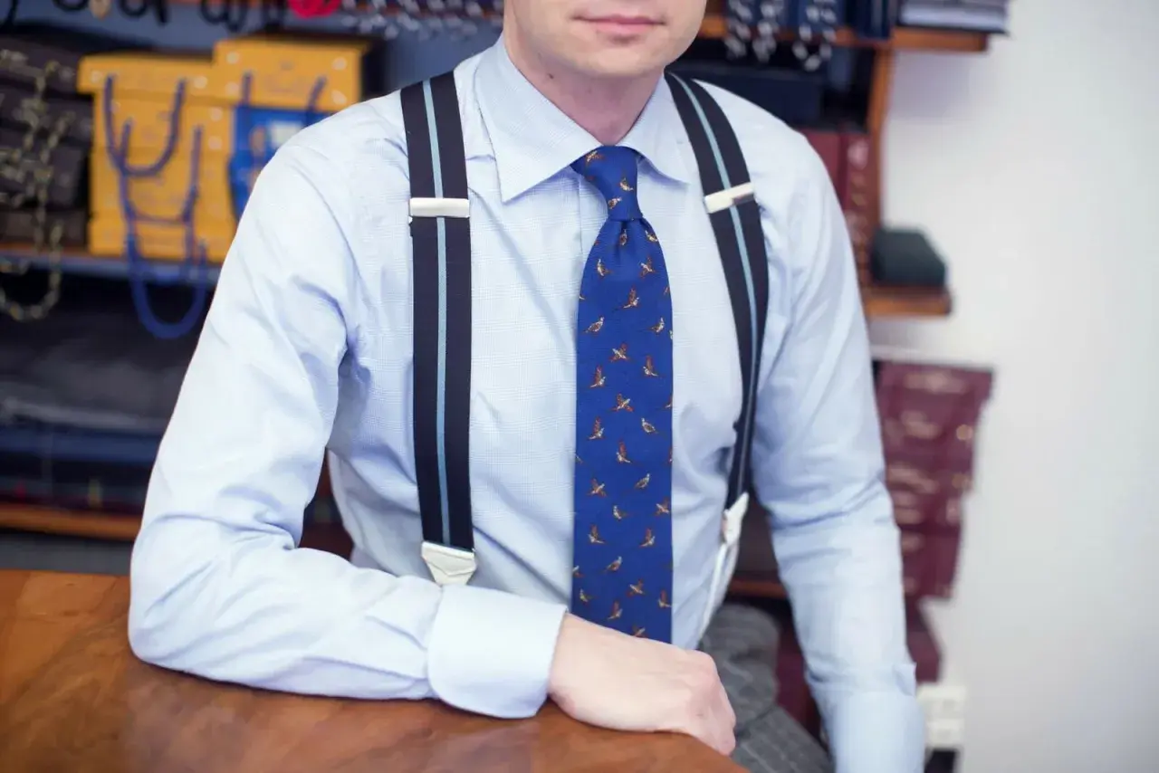 Elegancki mężczyzna w błękitnej koszuli i krawacie z ptakami, pokazuje, jak nosić szelki.