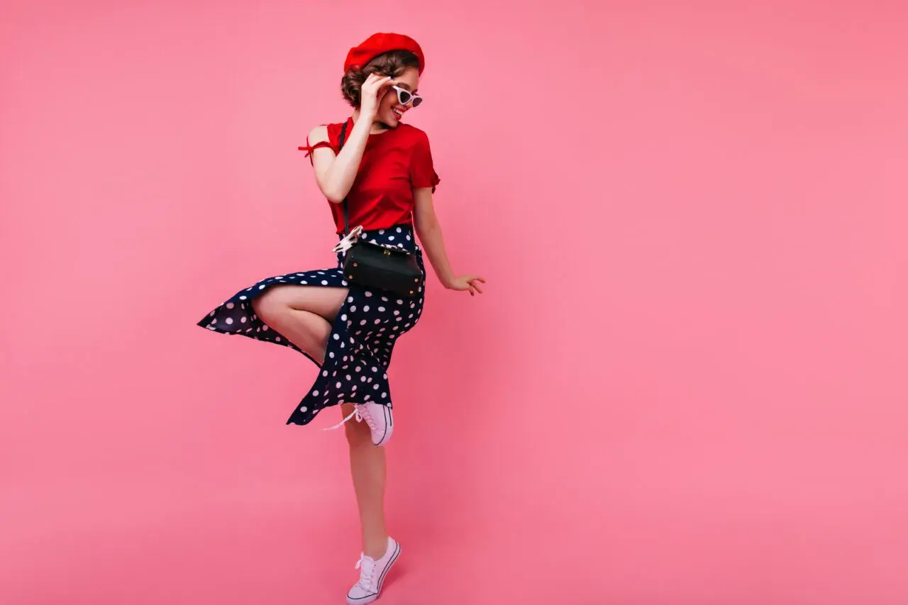 Dziewczyna w stylu pin-up girl w czerwonej bluzce, spódnicy w grochy i czerwonym beretcie skacze na różowym tle.
