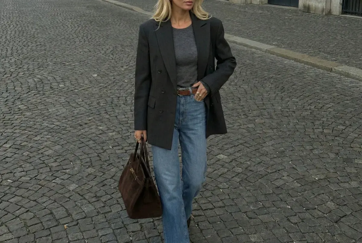 Elegancka kobieta w szarym longsleeve, jeansach i marynarce idzie po bruku, niosąc brązową torebkę.
