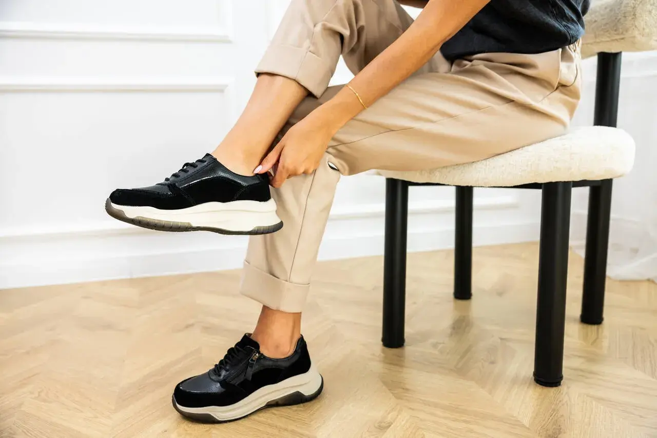Kobieta w beżowych spodniach i czarnych sneakersach na grubej podeszwie siedzi na krześle.