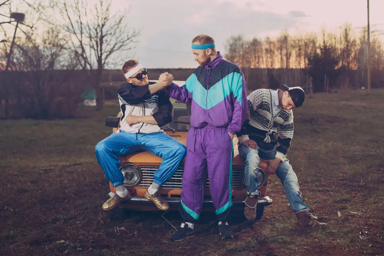 Trzech mężczyzn w dresach i okularach przeciwsłonecznych siedzi na masce starego samochodu. Stylizacja rodem z lat 90. xx wieku.