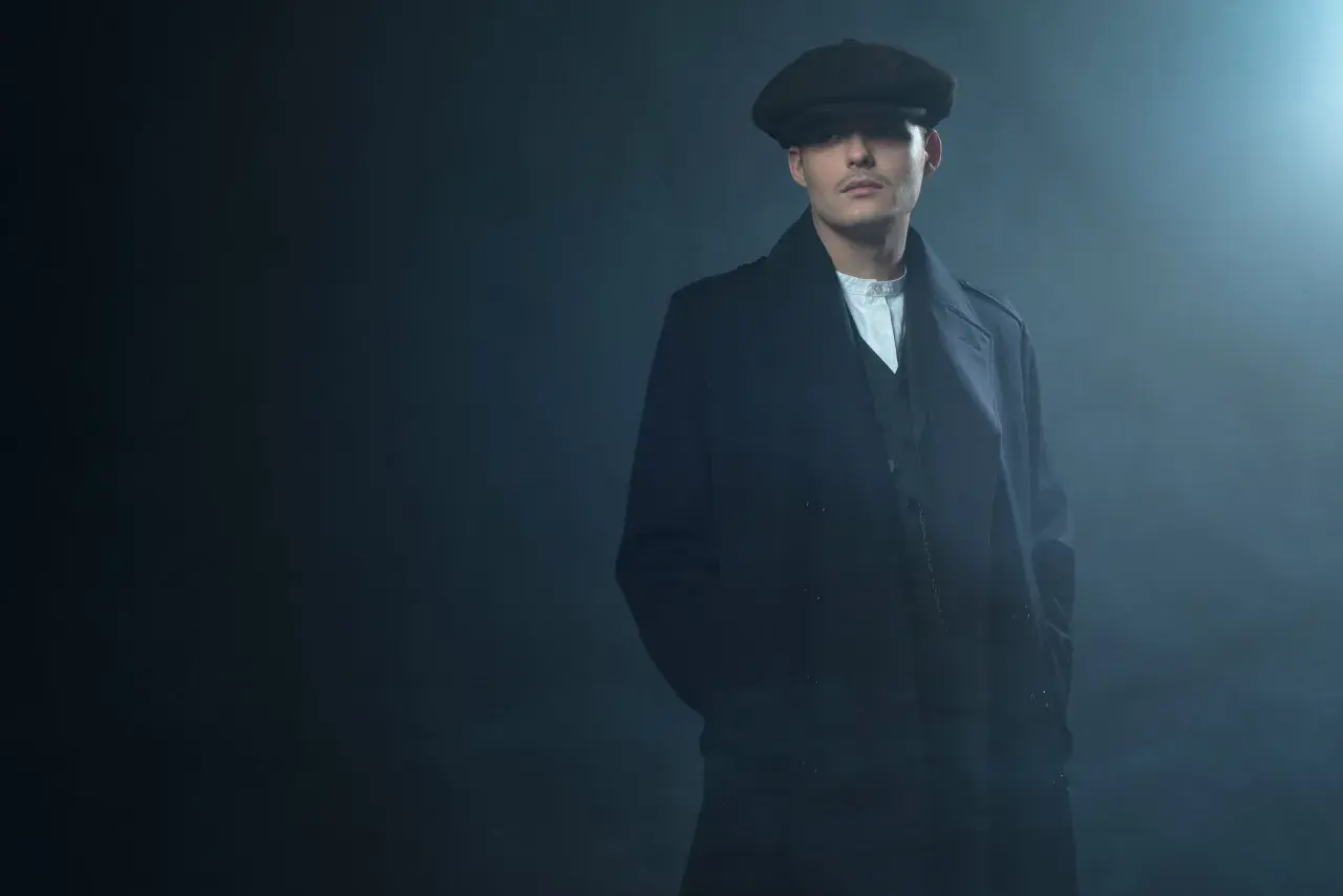 Mężczyzna w kaszkiecie i płaszczu, styl Peaky Blinders, z rękami w kieszeniach, w zadymionym otoczeniu.