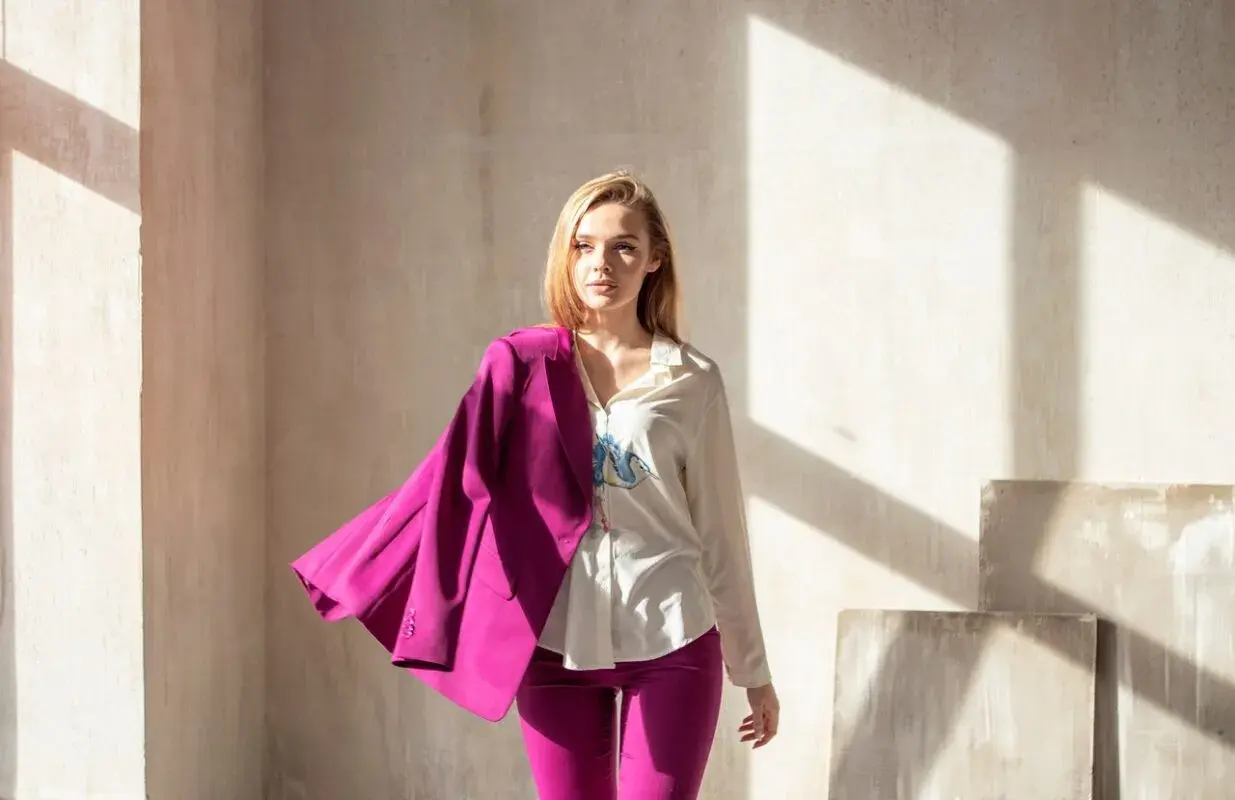Kobieta w stylizacji viva magenta, z marynarką narzuconą na ramię, stoi w świetle słonecznym.