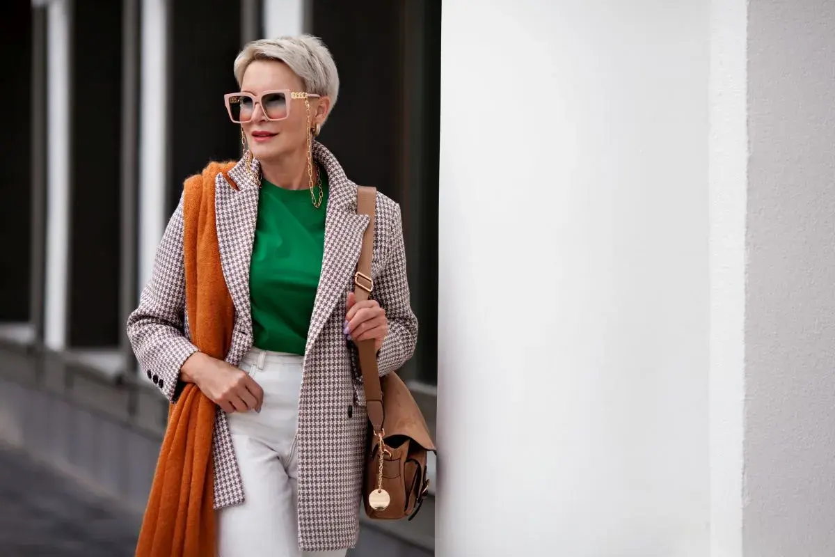 Elegancka kobieta po 60. w stylowej marynarce, zielonej bluzce i białych spodniach. Pomarańczowy szal dodaje jej uroku.