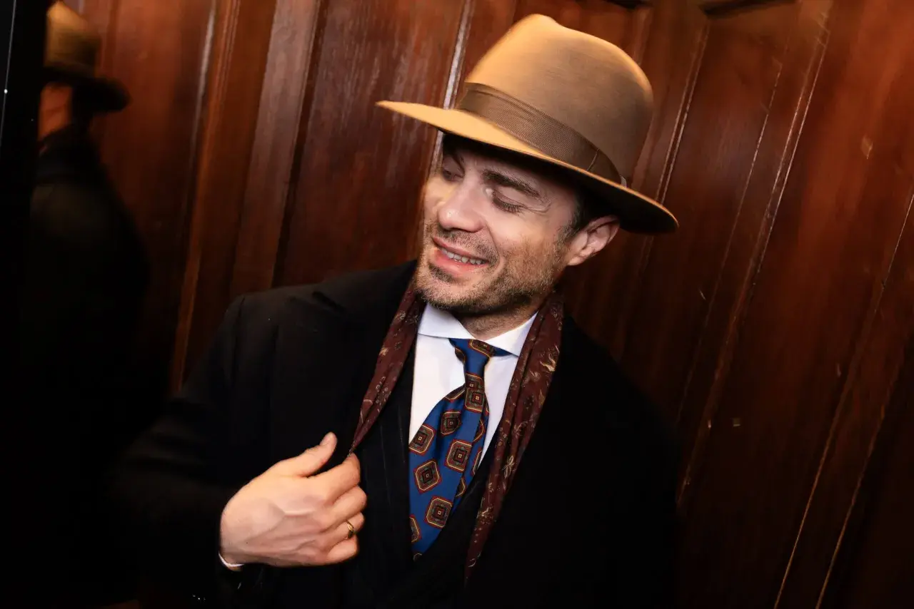 Elegancki mężczyzna w brązowym kapeluszu, czarnym płaszczu i stylowym krawacie. Pokazuje, jak nosić kapelusz męski, dodając klasy każdej stylizacji.