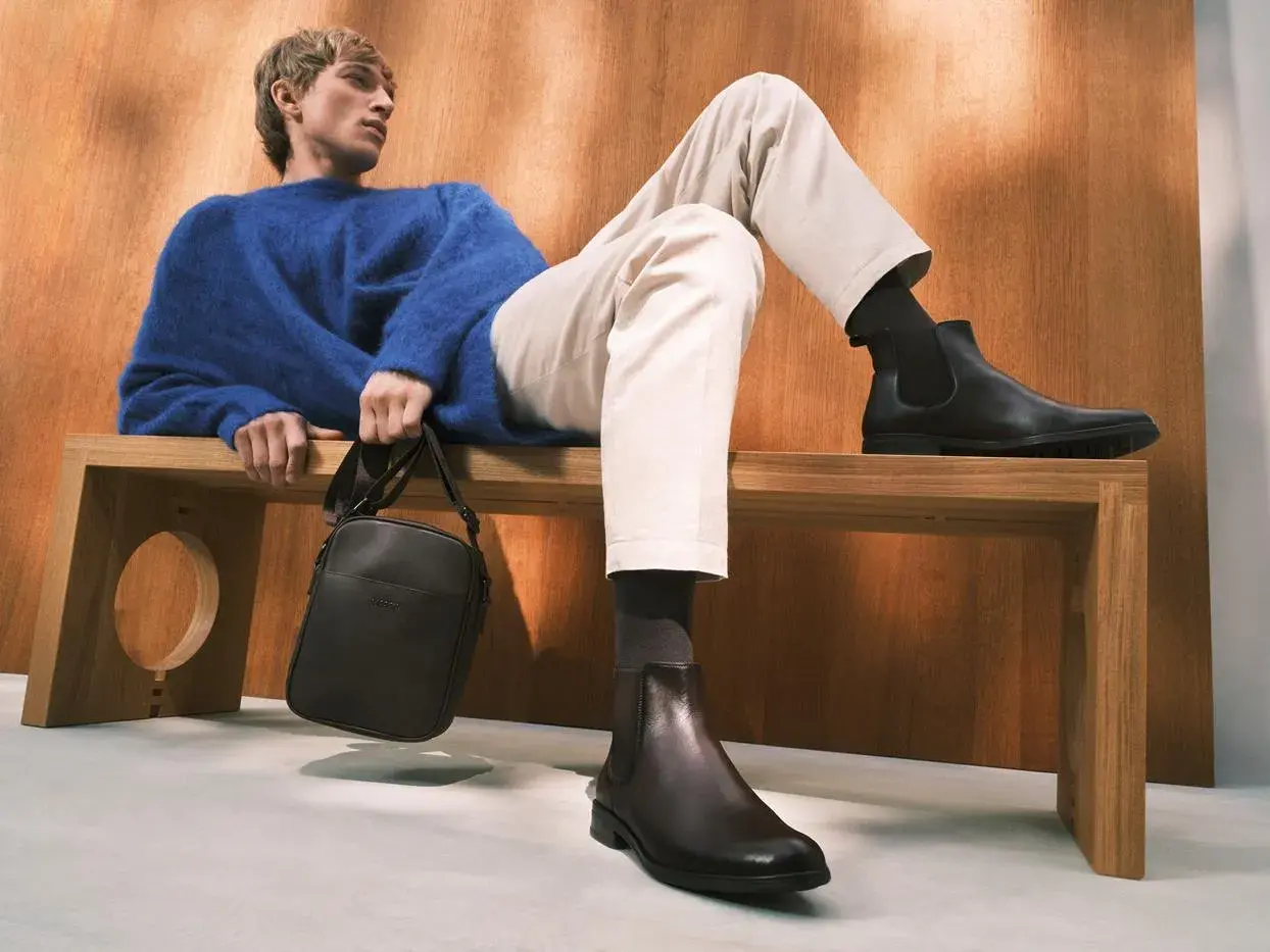 Eleganckie, ciemne botki to idealne buty do chinosów i koszuli. Mężczyzna w niebieskim swetrze prezentuje stylowy zestaw.