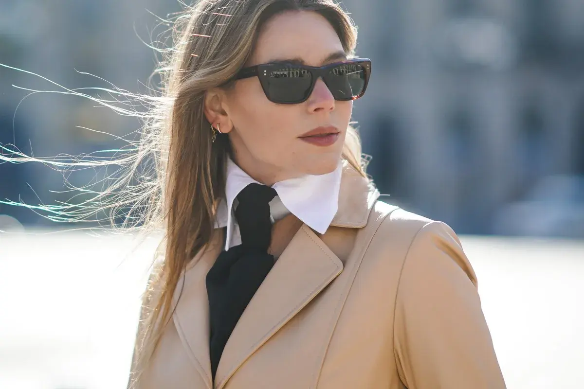 Elegancka kobieta w beżowym płaszczu, okularach przeciwsłonecznych i białej koszuli z czarną apaszką, tworzącą ciekawy rodzaj kołnierzyka.