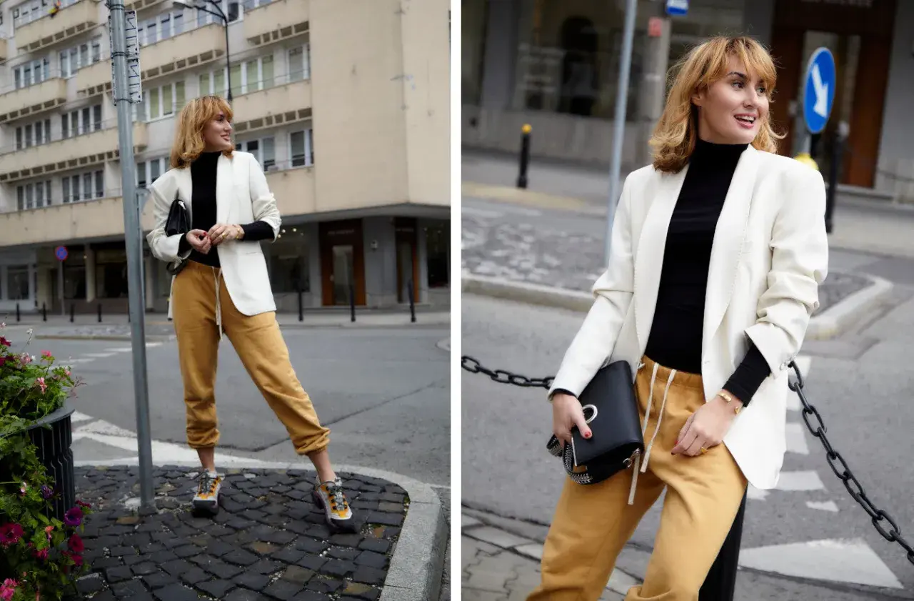 Modna kobieta w stylizacji z czarnym golfem, białej marynarce i musztardowych spodniach dresowych.
