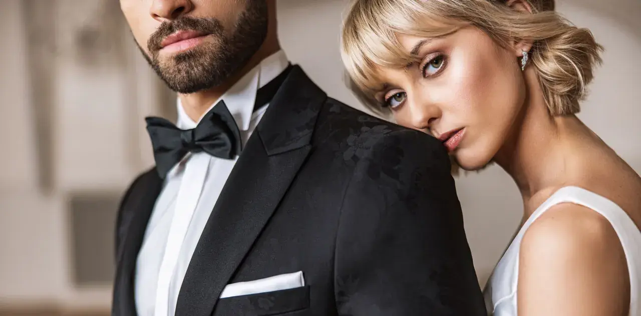 Elegancki mężczyzna w czarnym smokingu z muchą i białą koszulą. Kobieta opiera głowę na jego ramieniu. Idealny przykład, jaka koszula do muchy.