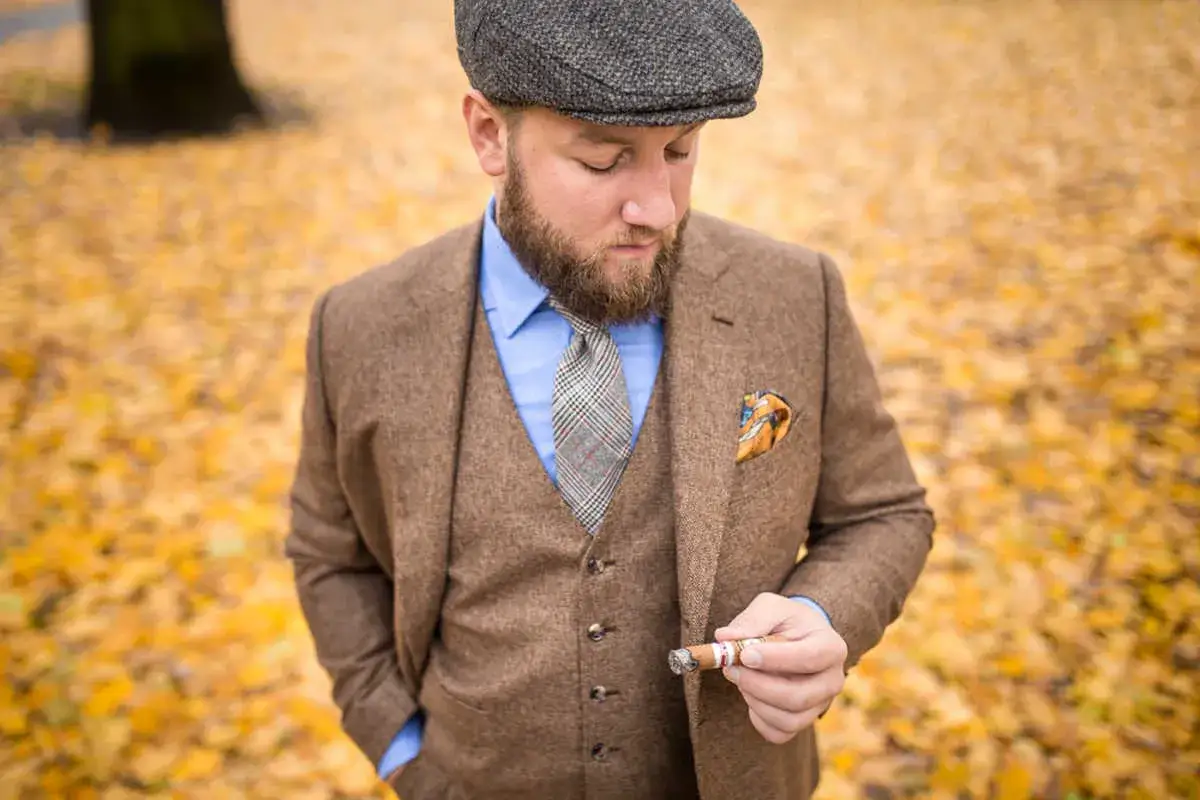 Elegancki mężczyzna w kaszkiecie i brązowym garniturze, trzymający cygaro. Stylowe kaszkiet męski stylizacje na jesień.