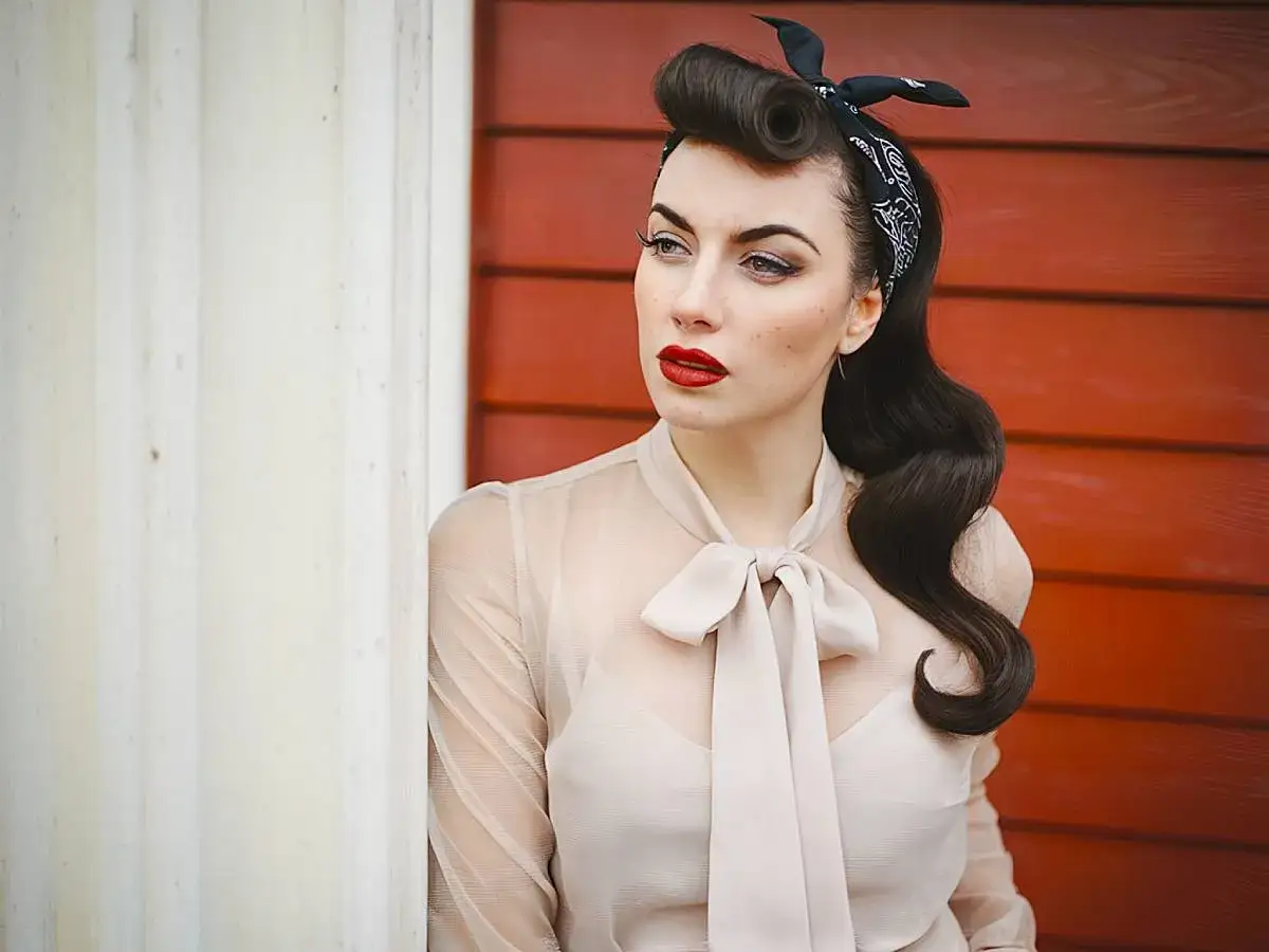 Stylowa pin-up girl z burzą ciemnych loków i czerwoną szminką, ubrana w transparentną bluzkę z kokardą, pozuje na tle czerwonej ściany.