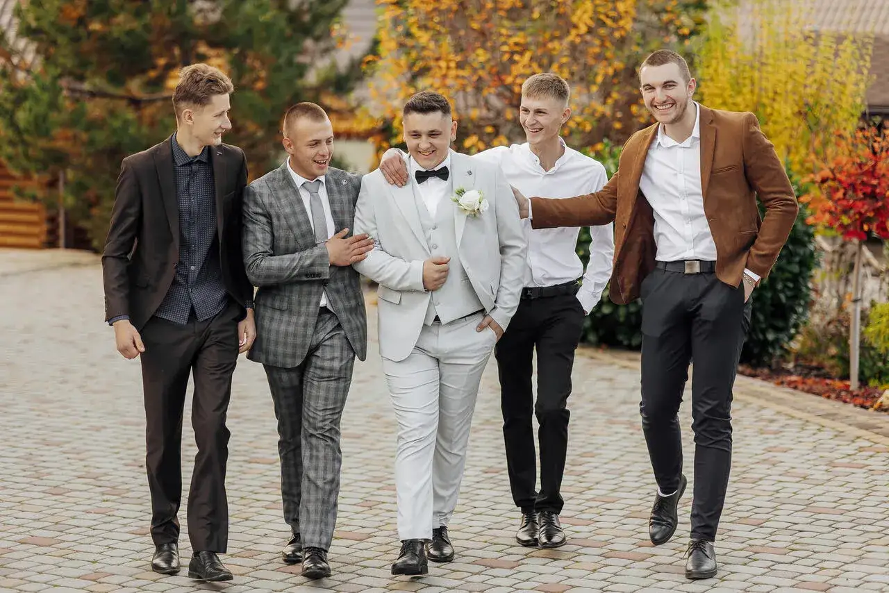 Pięciu mężczyzn w eleganckich strojach, w tym garniturach i marynarkach, idzie razem, ciesząc się towarzystwem.