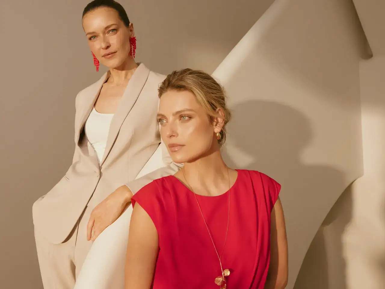 Dwie kobiety w eleganckich strojach, uosabiające styl power dressing. Jedna w beżowym garniturze, druga w czerwonej sukience.