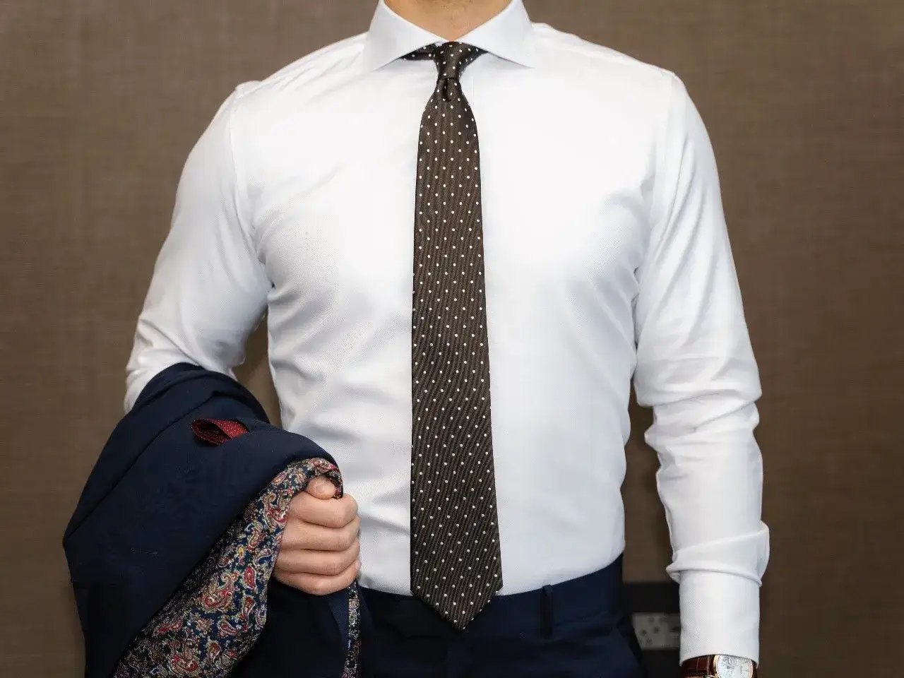 Elegancki mężczyzna w białej koszuli i brązowym krawacie w kropki. Jak wygląda krawat? Wzorzysta poszetka i granatowa marynarka dopełniają stylizację.