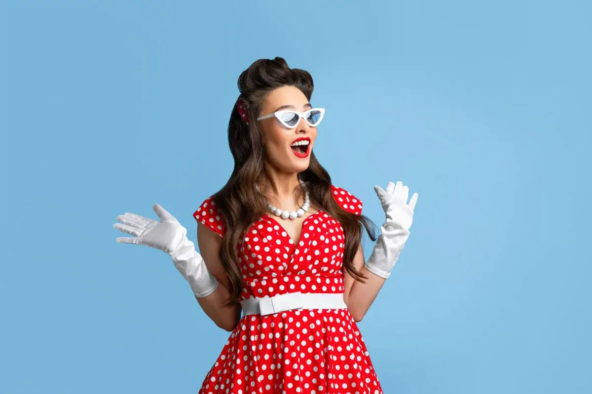 Kobieta w stylu pin-up w czerwonej sukience w grochy, białych rękawiczkach i okularach, z uniesionymi rękami.