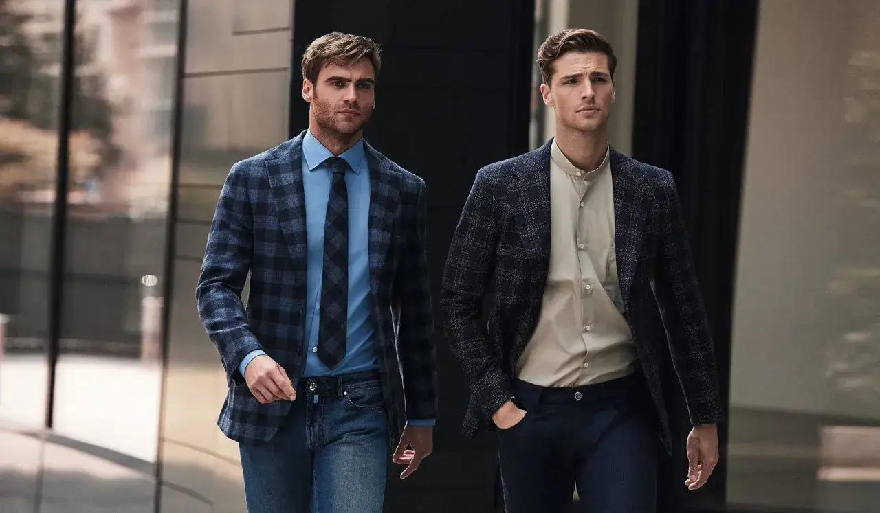 Dwóch mężczyzn w eleganckim stroju business casual, jeden w niebieskiej kratce i dżinsach, drugi w granatowej marynarce i beżowej koszuli.